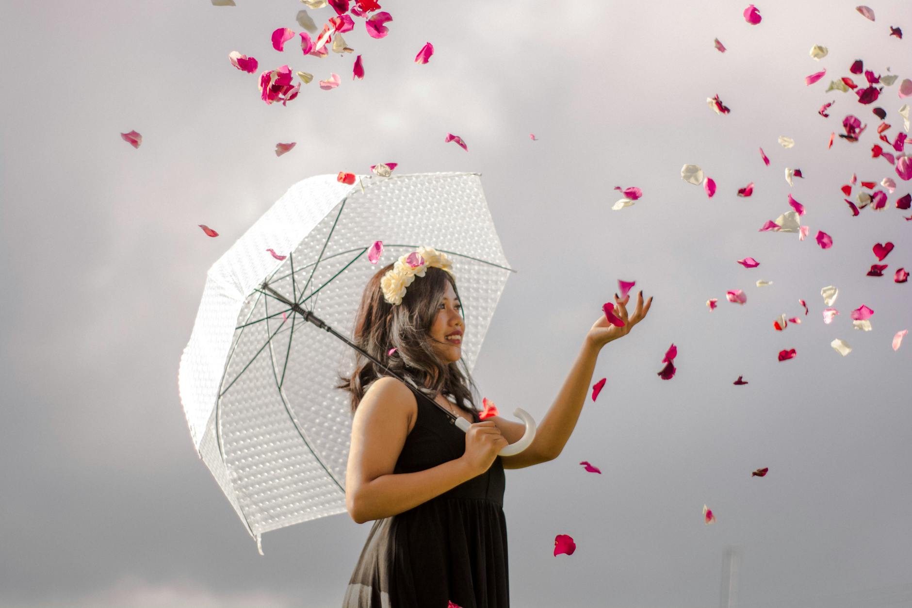 positive young ethnic woman throwing flower petals against cloudy sky